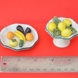 Puede incluir: Dos platos de cerámica con diseños de frutas. Un plato es blanco con hojas verdes y limones amarillos. El otro plato es blanco con un borde verde y presenta un plátano, dos naranjas, un tomate y dos berenjenas.