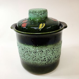 Könnte beinhalten: Eine grüne Keramikdose mit Deckel, mit einem gesprenkelten Überzug und einem bemalten Design aus Früchten und Blättern. Die Dose hat eine breite Öffnung und einen runden Körper.