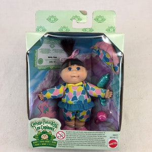 Op de afbeelding: Een Cabbage Patch Kids pop genaamd Molly Jean, gekleed in een blauw-roze geprint pakje, wordt geleverd met een roze-groene speen en een roze deken. De pop is verpakt in een doorzichtige plastic doos met een groene en witte achtergrond. De doos bevat ook een geboortecertificaat voor de pop, waarop staat dat ze op 15 juli is geboren. De doos is gelabeld met het Cabbage Patch Kids logo en de woorden "Les Copinoux".
