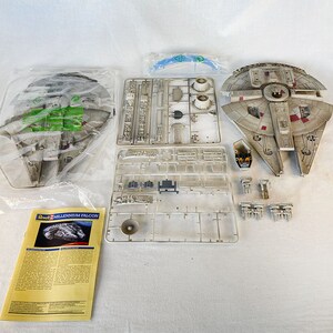 Puede incluir: Un kit de maquetas del  Millennium Falcon, la nave espacial de Star Wars. El kit incluye la nave, las instrucciones y varias piezas de plástico. La nave es gris y blanca con detalles rojos.