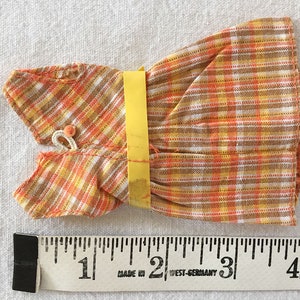 May include: A small orange, yellow, and white plaid dress with a yellow tag. The dress is about 4 inches long and is made in West Germany.