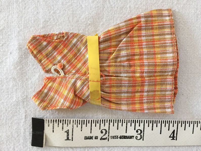 May include: A small orange, yellow, and white plaid dress with a yellow tag. The dress is about 4 inches long and is made in West Germany.