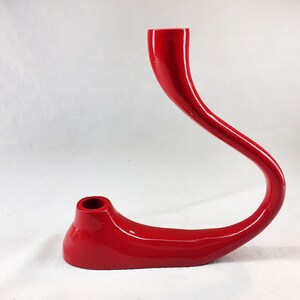 May include: A red, abstract, ceramic candle holder with a curved, elongated design. The holder has a smooth, glossy finish.