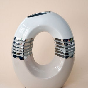 May include: White ceramic vase with a silver ring design. The vase has a unique oval shape with a hole in the center.