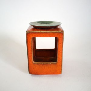 May include: A square, orange ceramic candle holder with a rectangular opening in the center. A small, round, green dish sits on top. The candle holder has a glossy finish and is set against a white background.