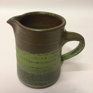 May include: A small, brown and green ceramic pitcher with a handle. The pitcher has a brown top and a green bottom.