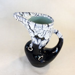 May include: A black and white ceramic pitcher with a crackled glaze. The pitcher has a unique, abstract shape with a spout and a handle. The bottom of the pitcher is marked with the word "Millau".