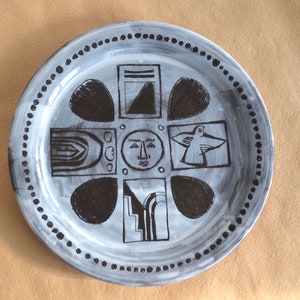 May include: A white ceramic plate with a black and white design. The design features a face, a bird, and geometric shapes.