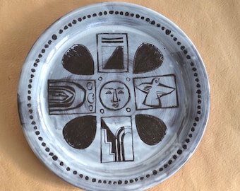 May include: A white ceramic plate with a black and white design. The design features a face, a bird, and geometric shapes.