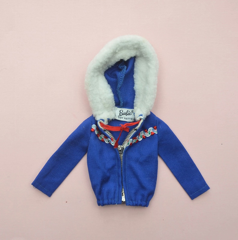 Op de afbeelding: Blauwe capuchontrui met een witte imitatiebontvoering en een rood lint. De trui heeft een ritssluiting en een witte en rode rand rond de capuchon. Het label luidt "Barbie by Mattel".
