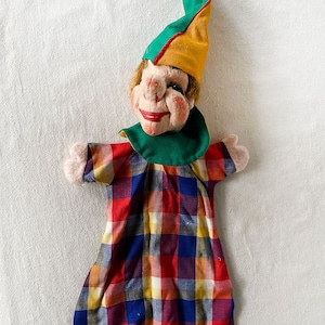 Puede incluir: Un títere de mano vintage con un sombrero verde y amarillo, un vestido de cuadros rojos, amarillos, azules y blancos y una cara sonriente.
