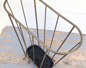 May include: A gold metal umbrella stand with a black metal base. The stand has a cylindrical shape and is made of thin metal bars.