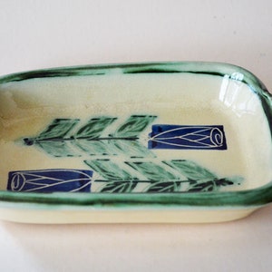 Puede incluir: Un plato rectangular de cerámica con un esmalte amarillo claro y diseños florales verdes y azules. El plato tiene dos asas a los lados.