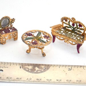 Op de afbeelding: Een miniatuur poppenhuis meubelset met een gouden kaptafel met spiegel, een tafel met een bloemenmotief en een bank met een bloemenmotief. De meubels zijn gemaakt van metaal en hebben een geverfde afwerking.