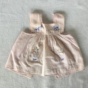 May include: A pink sleeveless dress with embroidered flowers and blue bows. The dress is made of a lightweight fabric and has a gathered skirt.