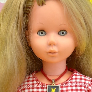 May include: A blonde doll with blue eyes wearing a red and white gingham dress and a red cord necklace with a silver pendant that has the letter "M" in yellow.