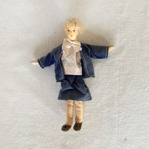May include: A wooden doll with blonde hair, wearing a blue jacket, a pink shirt with a bow, a blue skirt, and tan stockings. The doll is standing on a white surface.