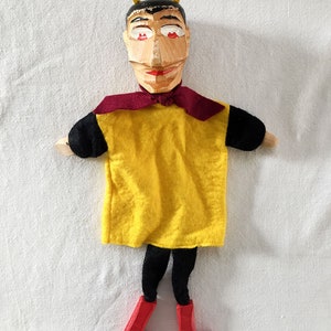 May include: A wooden hand puppet with a yellow shirt, black pants, and red shoes. The puppet has a carved face with horns, red eyes, and a red mouth.