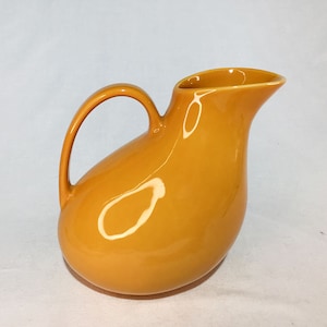 Könnte beinhalten: Eine glänzende, senfgelbe Keramikkrug mit einem gebogenen Griff und einem Ausguss. Der Krug hat einen abgerundeten Körper und eine glatte, reflektierende Oberfläche. Der Griff ist gewölbt und der Ausguss ist zum einfachen Ausgießen abgewinkelt.