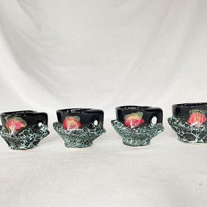Puede incluir: Cuatro tazas de cerámica negra con un esmalte moteado verde y rojo. Las tazas tienen una forma única y orgánica con un asa en el lateral.