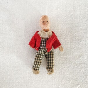 May include: A small cloth doll with a red jacket, black and white checkered pants, and a light brown head. The doll has painted eyes and a simple smile.