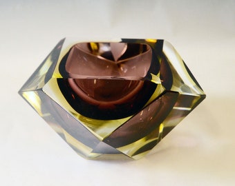 Puede incluir: Un cenicero de vidrio geométrico con un interior morado oscuro y un exterior verde claro. El cenicero tiene un diseño facetado y está hecho de vidrio grueso.