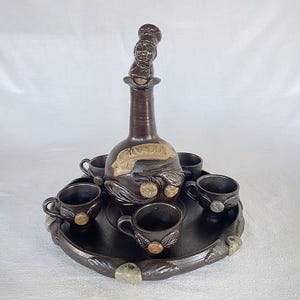 May include: A brown ceramic drinks set featuring a decanter with a chef figurine and six small cups on a round tray. The decanter has a decorative design with fruit and leaves. The cups and tray have a matching design. The set is made of ceramic.