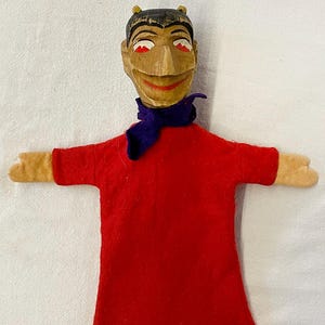 Puede incluir: Un títere de mano de madera con un cuerpo de fieltro rojo y una bufanda de fieltro morado. El títere tiene una cabeza de madera tallada con una cara pintada, cuernos y ojos rojos.