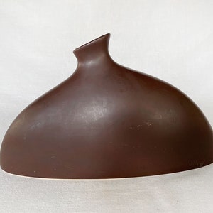 May include: A brown ceramic vase with a unique, asymmetrical shape. The vase has a wide, rounded base and a narrow, curved neck.