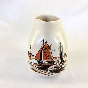 May include: A white ceramic vase with a brown and black sailboat design. The vase has a rounded shape and a slightly uneven opening.