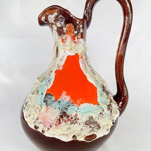 May include: A vintage ceramic pitcher with a dark brown base and handle. The pitcher features a vibrant orange center, accented by textured white, blue, and orange details. The handle curves gracefully from the top to the base, adding to its unique design.