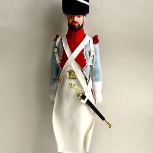 Puede incluir: Una figurita de soldado de juguete que lleva un uniforme azul y blanco con una faja roja y un sombrero de piel negro con una pluma roja. El soldado sostiene una espada y está de pie sobre una base negra con una inscripción dorada que dice "INFANTERIE DE LIGNE  SAPEUR 3 ème REGIMENT".