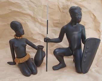 May include: Two black ceramic figurines of a man and woman. The man is kneeling and holding a spear, while the woman is sitting and holding a vase. Both figurines have beaded necklaces and are painted in a dark brown colour.