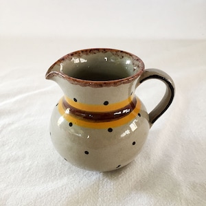May include: A small, cream-colored ceramic pitcher with a brown rim and handle. The pitcher is decorated with black polka dots and a yellow and brown stripe.