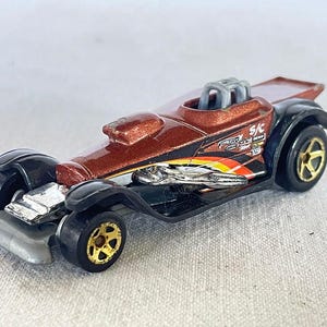 May include: A die-cast toy car in shades of brown, black, and silver. The car features gold-colored wheels and a detailed engine compartment. The side of the car has a graphic with the text "S/C".
