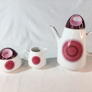 May include: A white ceramic tea set with a pink and burgundy design. The set includes a teapot, creamer, and sugar bowl.