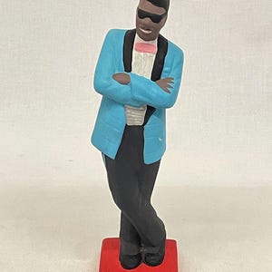 Könnte beinhalten: Keramikfigur eines Mannes in hellblauer Jacke, schwarzer Hose und Sonnenbrille. Die Figur hat verschränkte Arme und steht auf einem roten Sockel. Die Jacke hat schwarze Revers und eine rosa Fliege.