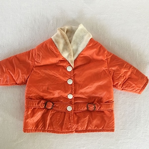 May include: An orange quilted jacket with a cream-colored ribbed collar and three white buttons. The jacket features a belt with two buckles at the waist. The jacket is a vintage style.