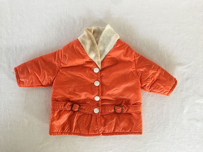 May include: An orange quilted jacket with a cream-colored ribbed collar and three white buttons. The jacket features a belt with two buckles at the waist. The jacket is a vintage style.