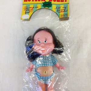 May include: A plastic doll with black hair, blue eyes, and a pink face. The doll is wearing a blue and white checkered top and matching bottoms. The doll is packaged in a plastic bag with a cardboard header that reads "Action Doll!" and "See my funny actions!"