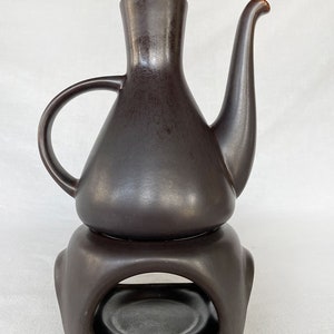 May include: A dark brown ceramic teapot with a spout and a handle, sitting on a matching ceramic stand with a circular opening. The stand has a small, round platform in the center.