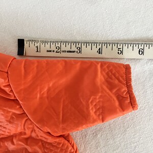 May include: Bright orange jacket sleeve with quilted texture, approximately 7 inches long. A wooden ruler with black markings is placed next to the sleeve for measurement. The jacket appears to be made of a shiny, synthetic material.