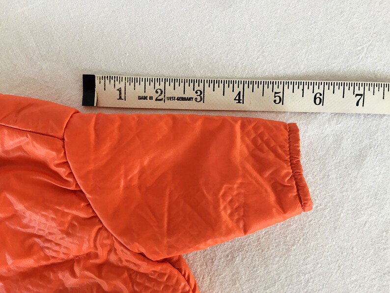 May include: Bright orange jacket sleeve with quilted texture, approximately 7 inches long. A wooden ruler with black markings is placed next to the sleeve for measurement. The jacket appears to be made of a shiny, synthetic material.