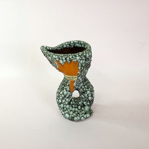 May include: A green and orange ceramic planter with a textured surface. The planter has a unique shape and a hole in the side. The planter has a label that reads "Danish Design".