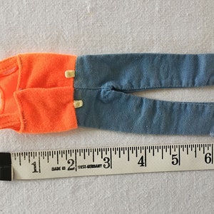 May include: A pair of doll clothes, a sleeveless orange shirt and blue jeans. The clothes are laid out on a white surface with a ruler for scale. The ruler is marked in inches.