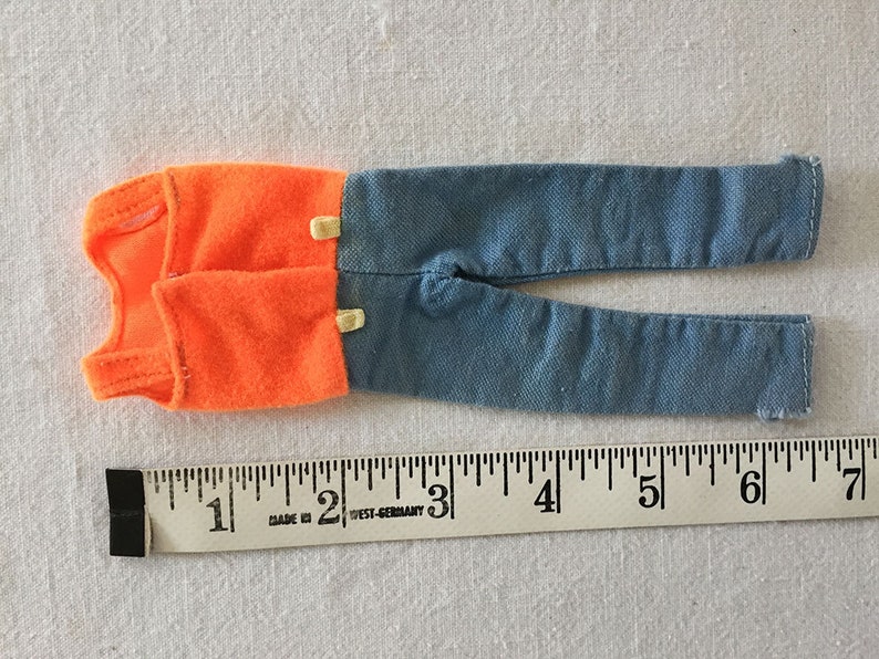 May include: A pair of doll clothes, a sleeveless orange shirt and blue jeans. The clothes are laid out on a white surface with a ruler for scale. The ruler is marked in inches.