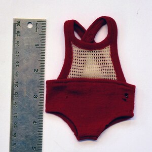 Op de afbeelding: Een bordeauxrode poppenromper met een crèmekleurig mesh paneel. De romper heeft een kruisbandontwerp en is ongeveer 7.6 cm lang. Een metalen liniaal met inch-markeringen ligt naast de romper.
