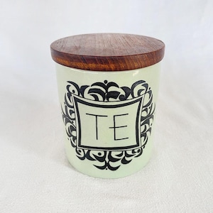 May include: A light green ceramic tea canister with a wooden lid. The canister features a black decorative frame with the word "TE" in the center. The tea container is designed for storing tea leaves.