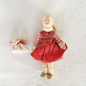 May include: A handmade doll with a red skirt and a checkered top, alongside a small fabric bag with a red and white checkered ribbon. The doll has blonde hair and is wearing a white collar. The bag has a pattern of pink hearts.