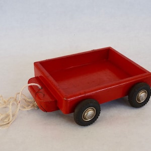 Puede incluir: Un carro de juguete de madera rojo con cuatro ruedas y una cuerda atada a la parte trasera. El carro tiene un diseño sencillo y está hecho de madera sin terminar.